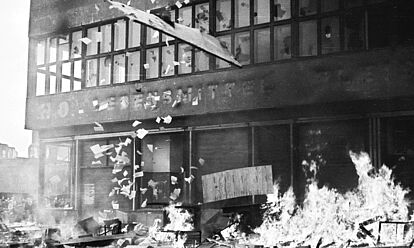 Das Schwarz-Weiß-Bild zeigt ein Feuer vor dem Columbia-Haus am Potsdamer Platz in Berlin. Aus dem Haus fliegen Papiere und ein fenster. Die meisten fensterscheiben des Hauses sind beschädigt.