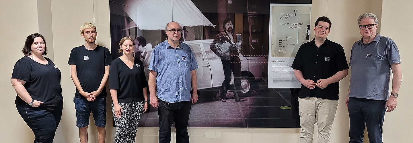 Sechs Personen stehen vor einer Wand, auf der ein großformatiges Foto von Wolf Biermann und vom Aktendeckel des Zentralen Operativen Vorgangs 'Lyriker' zu sehen ist.