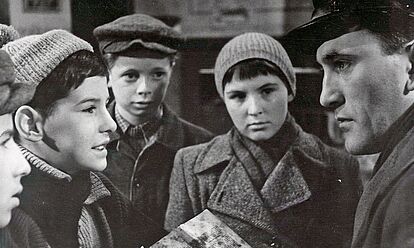 Das Bild zeigt eine Szene aus dem Film „Die Jagd nach dem Stiefel' von 1962. Eine Gruppe von Kindern in Winterkleidung unerhalten sich mit einem erwachsenen Mann, der ein Buch in der Hand hat und sich zu den Kindern heruntergebeugt hat.