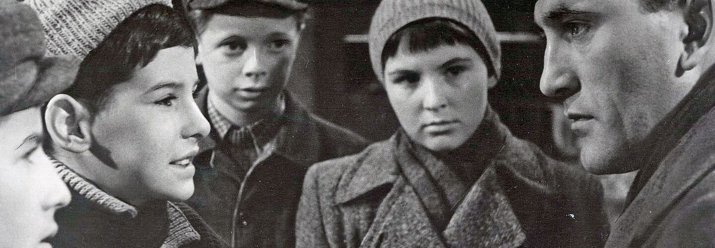 Das Bild zeigt eine Szene aus dem Film „Die Jagd nach dem Stiefel' von 1962. Eine Gruppe von Kindern in Winterkleidung unerhalten sich mit einem erwachsenen Mann, der ein Buch in der Hand hat und sich zu den Kindern heruntergebeugt hat.