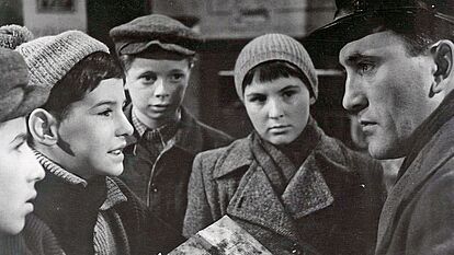 Das Bild zeigt eine Szene aus dem Film „Die Jagd nach dem Stiefel' von 1962. Eine Gruppe von Kindern in Winterkleidung unerhalten sich mit einem erwachsenen Mann, der ein Buch in der Hand hat und sich zu den Kindern heruntergebeugt hat.