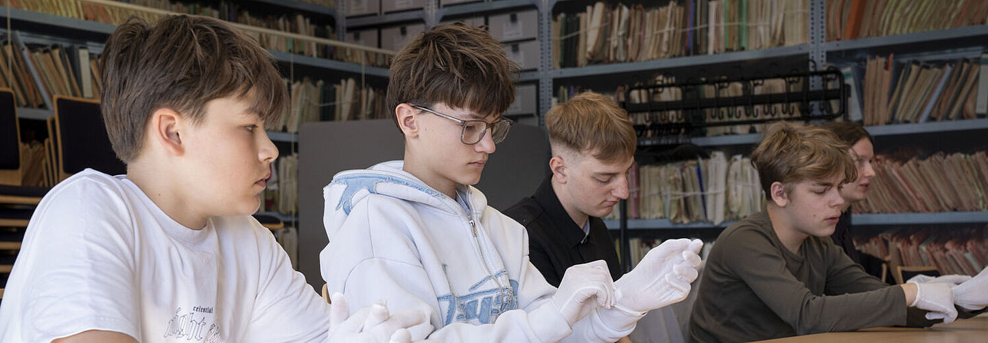 Eine Gruppe von fünf Jungen sitzt nebeneinander an einem Tisch. Die Jungen ziehen weiße Handschuhe an.
