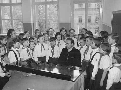 Eine Gruppe von Kindern steht neben einem Klavierflüge, an dem ein Mann sitzt. Alle Personen singen.