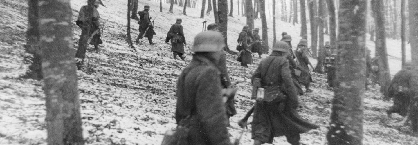 Deutsche Soldaten laufen durch einen verschneiten Wald. Im Vordergrund ist ein Soldat mit einem Sturmgewehr 44 zu sehen.