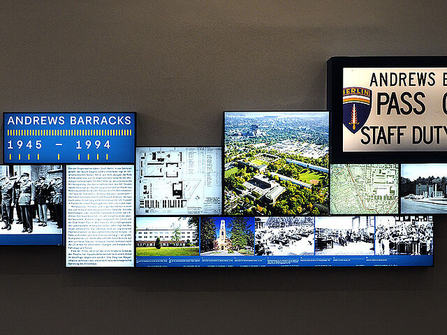 Beleuchtete Fotos und Texte sowie eine Vitrine mit einem Schild der Andrews Barracks sind an eine Wand montiert.