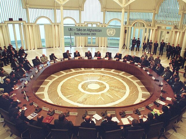 Mehrere Frauen und Männer sitzen an einem runden Tisch. Im Hintergrund hängt ein Banner mit der Aufschrift „UN-Talks on Afghanistan. Bonn, November 2001".