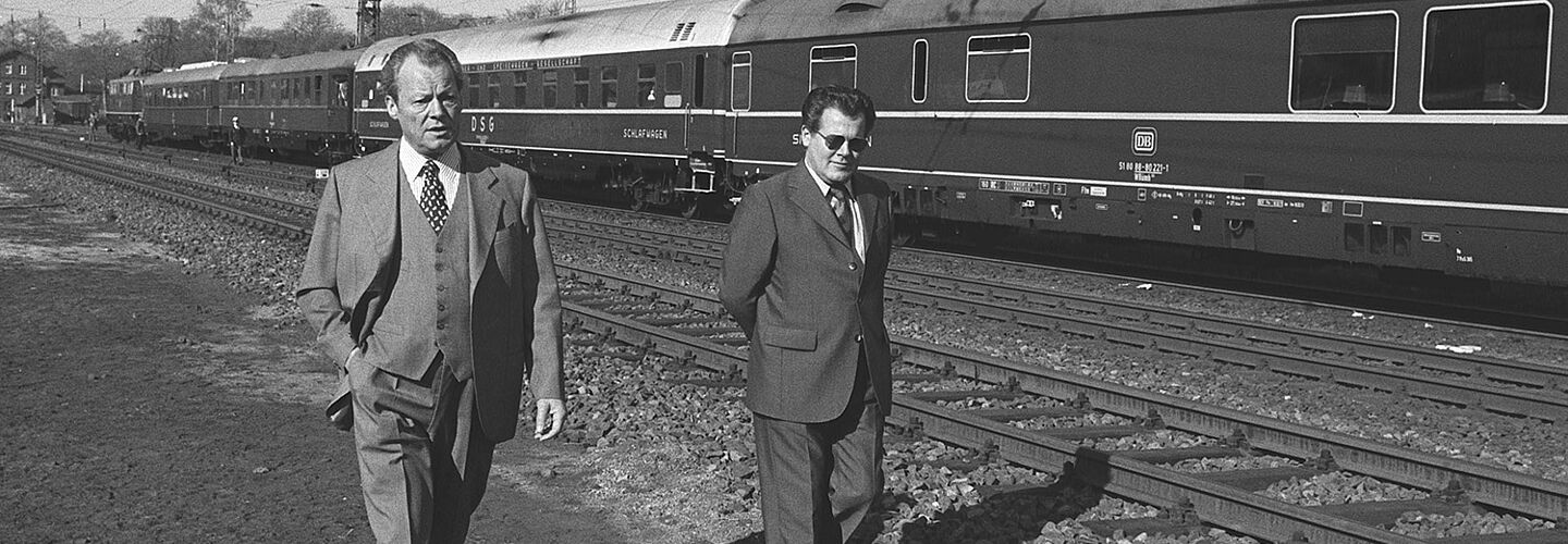 Schwarz-Weiß-Aufnahme mit Willy Brandt und Günter Guillaume, bekleidet mit Anzug und Krawatte und an Bahngleisen entlanggehend, im Hintergrund ein Personenzug