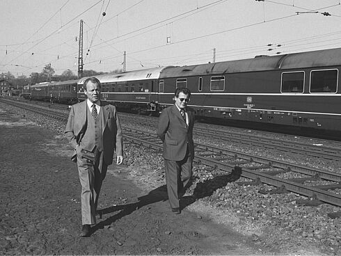 Schwarz-Weiß-Aufnahme mit Willy Brandt und Günter Guillaume, bekleidet mit Anzug und Krawatte und an Bahngleisen entlanggehend, im Hintergrund ein Personenzug