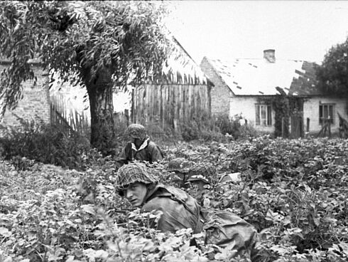 Soldaten der Waffen-SS begeben sich in Sträuchern in Deckung. Im Hintergrund sind ein Baum, ein Zaun und ein Haus zu sehen.
