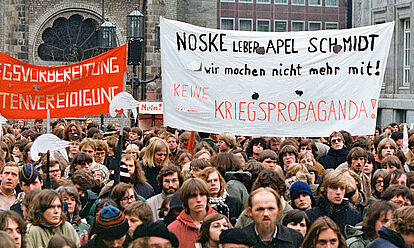 Aus einer großen Gruppe von Demonstranten heraus wird während einer Demonstration gegen ein öffentliches Gelöbnis der Bundeswehr in Bonn ein Transparent mit der Aufschrift „Noske Leber Apel Schmidt wir machen nicht mehr mit! Keine Kriegspropaganda!“ hochgehalten.