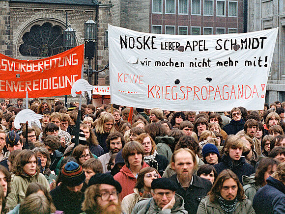 Aus einer großen Gruppe von Demonstranten heraus wird während einer Demonstration gegen ein öffentliches Gelöbnis der Bundeswehr in Bonn ein Transparent mit der Aufschrift „Noske Leber Apel Schmidt wir machen nicht mehr mit! Keine Kriegspropaganda!“ hochgehalten.