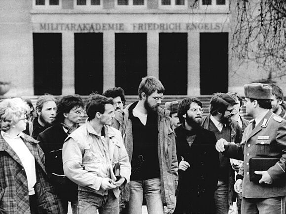 Zivilistinnen und Zivilisten in Alltagskleidung besuchen im Januar 1990 die Militärakademie „Friedrich Engels“ in Dresden. Es handelt sich um Vertreterinnen und Vertreter basisdemokratischer Gruppen aus Dresden. Die Gruppe steht vor dem Eingang, über dem der Schriftzug „Militärakademie ‚Friedrich Engels‘“ angebracht ist. Rechts im Bild steht ein Soldat in Uniform, der mit den Besucherinnen und Besuchern spricht.
