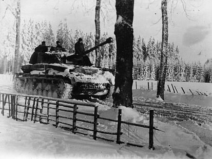 Ein schneebedeckter Panzer, auf dem Soldaten sitzen, fährt eine verschneite Straße entlang. Im Hintergrund ist ein Wald zu sehen.