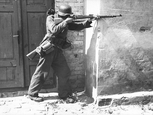 Ein deutscher Soldat steht mit einem Gewehr (Karabiner K 98) an einer Hausecke und zielt.