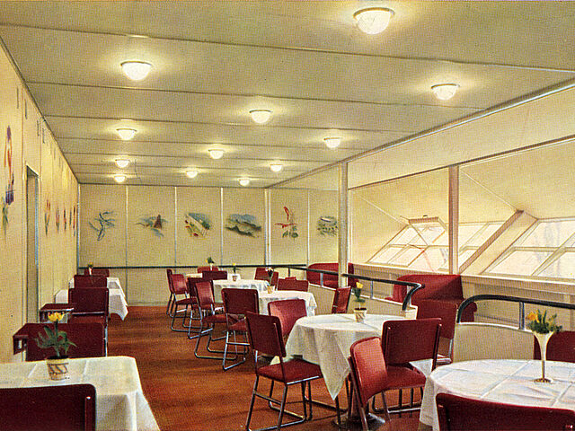 Blick in den leeren Speisesaal im Luftschiff LZ 129 „Hindenburg“. Zu sehen sind acht Tische mit weißen Tischdecken sowie rote Stühle.
