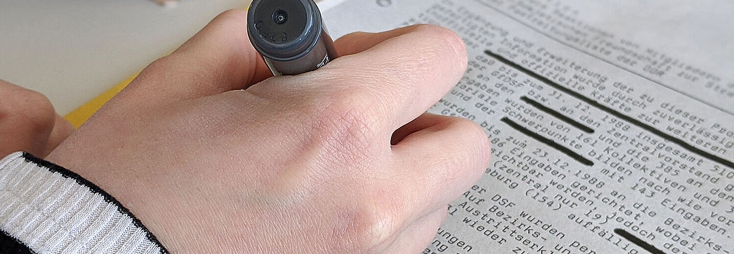 The image shows the hand of a female employee using a black pen to redact a Stasi document.