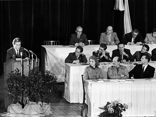 Links im Bild ist der Autor Werner Bräunig am Rednerpult zu sehen, auf der rechten Seite sitzen an Tischen Autorinnen und Autoren, die sich Bräunig zuwenden.