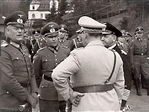 Es handelt sich um eine Schwarz-Weiß-Aufnahme von vier Herren in Uniformen, die miteinander reden. Im Hintergrund sind viele weitere uniformierte Männer sowie ein mit Bäumen bewachsener Hang und ein großes Gebäude zu sehen.