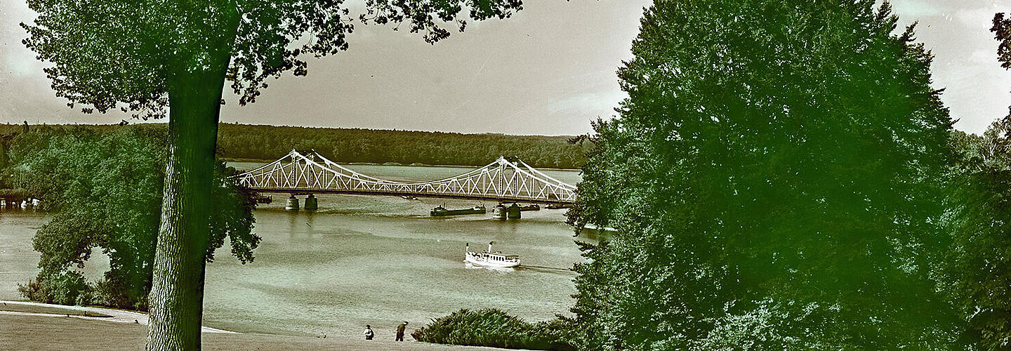 Die Farbfotografie wurde vom Ufer der Havel am Potsdamer Park Babelsberg aus aufgenommen. Im Bildmittelpunkt ist die Glienicker Brücke zu sehen. Auf dem Wasser fahren Schiffe. Hinter der Brücke ist ein Wald zu erkennen. Am Ufer im Bildvordergrund sind Bäume, Sträucher und zwei Personen zu sehen.