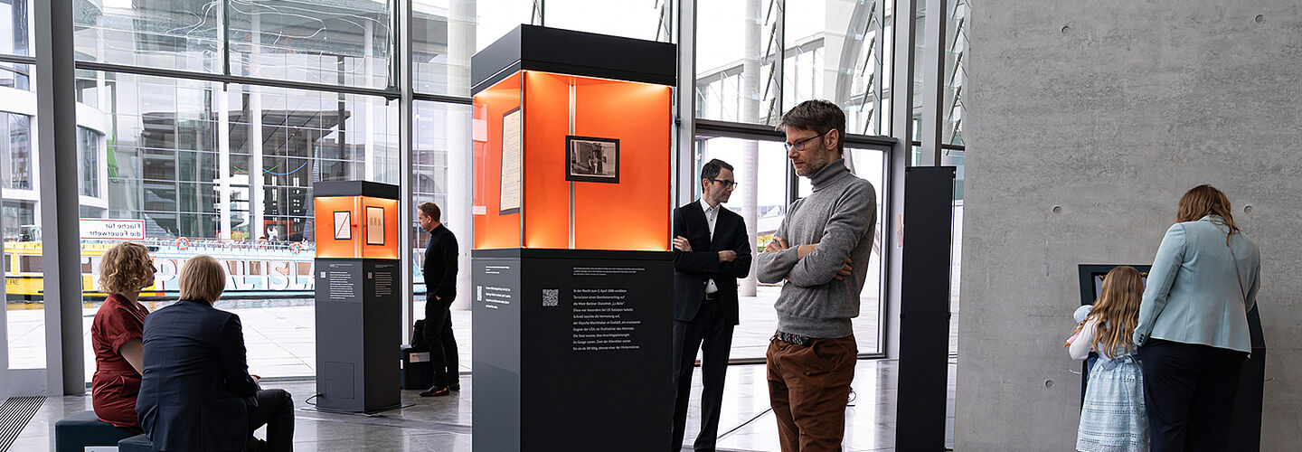 Ausstellungsstelen verteilt in einem Raum mit Glasfront, Menschen betrachten die ausgestellten Objekte