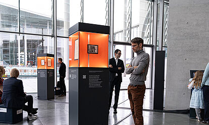 Ausstellungsstelen verteilt in einem Raum mit Glasfront, Menschen betrachten die ausgestellten Objekte