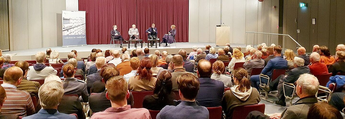 Ein gefüllter Saal ist zu sehen mit Blick auf die Bühne. Auf der Bühne sitzen vier Personen und halten Mikrofone in den Händen.