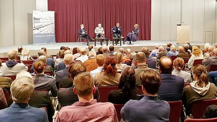 Ein gefüllter Saal ist zu sehen mit Blick auf die Bühne. Auf der Bühne sitzen vier Personen und halten Mikrofone in den Händen.
