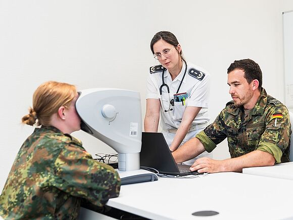 Eine Soldatin blickt in ein medizinisches Gerät. Vor ihr sitzt ein Soldat an einem Laptop. Nebem dem Mann steht eine Frau im Rang einer Sanitätsoffizierin. Sie trägt eine weiße Uniform.
