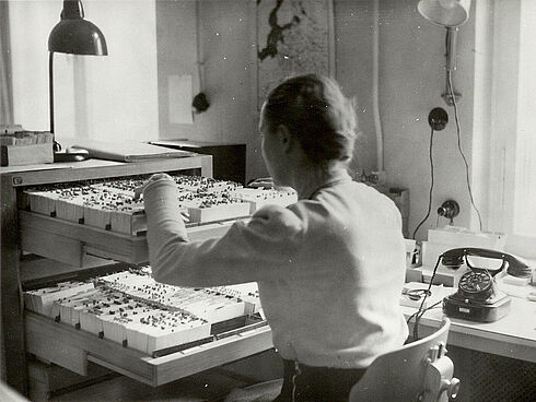 Schwarz-Weiß-Aufnahme mit einer Frau, die in einem Büro vor herausgezogenen Schubladen sitzt und in Karteikästen blättert