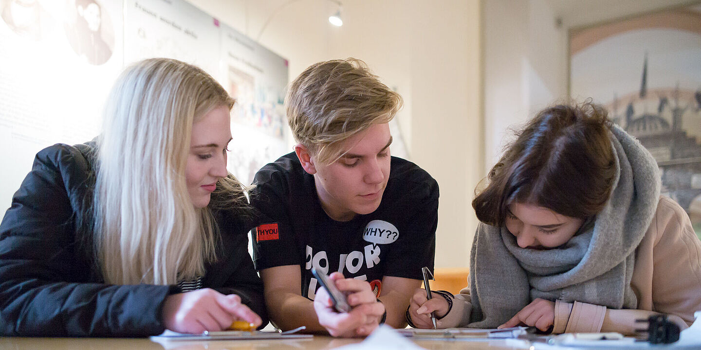 Drei Jugendliche sitzen an einem Tisch und vertiefen sich in eine schriftliche Aufgabenstellung., Quelle:
            Bundesarchiv