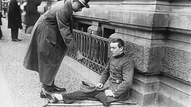 Ein einbeiniger Kriegsversehrter in Uniform mit Orden (Eisernes Kreuz I. Klasse, Verwundetenabzeichen) lehnt sitzend an einer Hauswand und bettelt. Ein Passant wirft Geld in die Mütze des Kriegsversehrten.