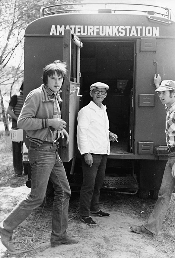 Drei Männer stehen vor einem Wagen mit der Aufschrift "Amateurfunkstation". 