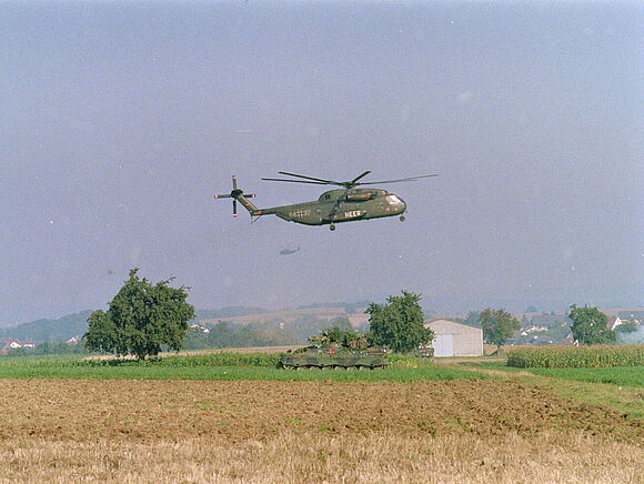 Ein Transporthubschrauber der Bundeswehr vom Typ Sikorsky CH-53 im Flug über einem abgeernteten Getreidefeld. Unter dem Hubschrauber steht ein getarnter Schützenpanzer „Marder“.