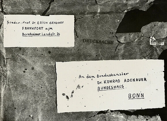 Aufgeklebtes Schwarz-Weiß-Foto eines mit Band verschnürten Pakets mit handgeschriebener Absender- und Empfängeradresse auf Etiketten, Briefmarken und „Drucksache“-Stempel; das Packpapier ist stark beschädigt.