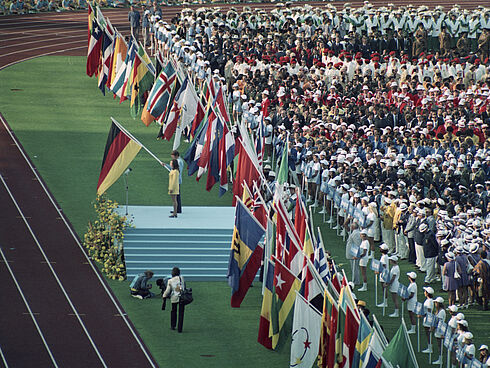 Farbfoto mit Fahnenträgern der Teilnehmerländer der Olympischen Sommerspiele 1972 in München. Heidi Schüller spricht auf einem Podest das Gelöbnis.