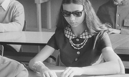 Auf dem Foto sieht man ein Mädchen, das an einem Tisch sitzt. Sie trägt eine Sonnenbrille und hat ihre Hände vor sich auf den Tisch gelegt. Im Hintergrund sieht man zwei weitere Personen.