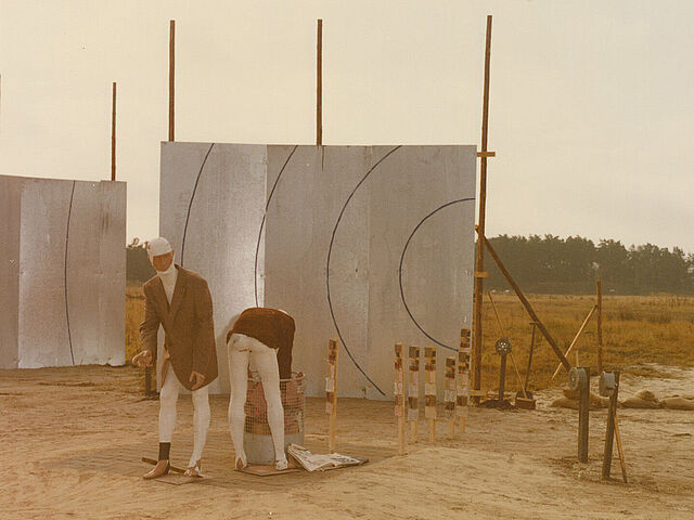 Zwei Puppen stehen vor zwei metallenen Wänden. Eine davon beugt sich über einen metallenen Mülleimer.