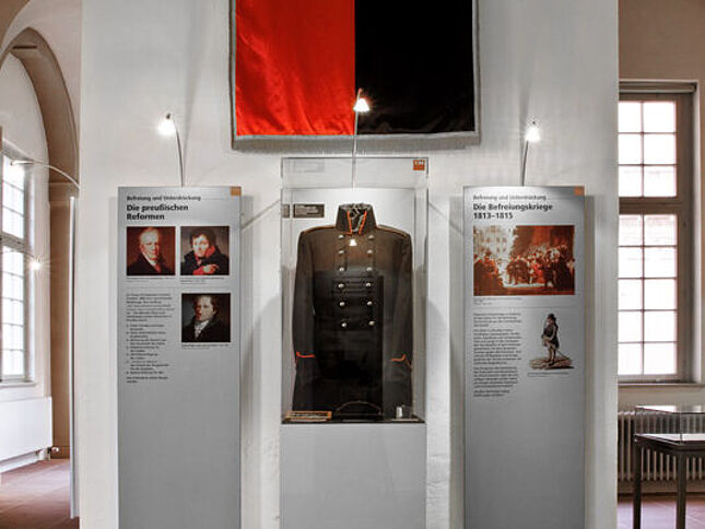 Die schwarz-rote Fahne der Jenenser Urburschenschaft hängt über einer Glasvitrine mit dem dunklen Uniformrock eines Lützower Jägers.