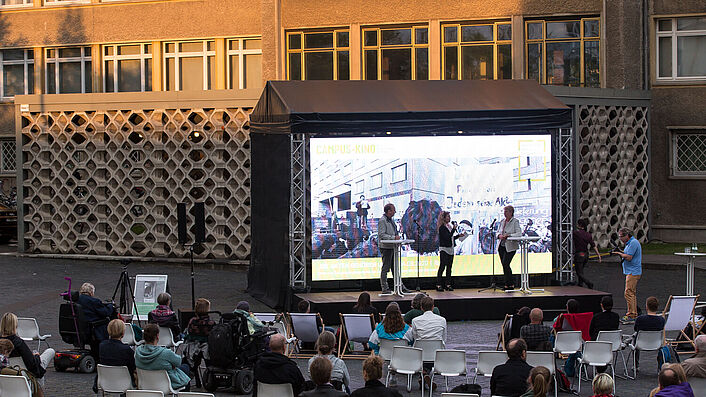 Diskussion auf einer Bühne, im Vordergrund sitzen Zuschauerinnen und Zuschauer auf Stühlen.