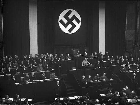Schwarz-Weiß-Aufnahme mit Adolf Hitler am Rednerpult im Reichstag, rundherum mit Männern in Anzügen vollbesetzte Sitzreihen; an der Hinterwand ein Banner mit Hakenkreuz-Symbol