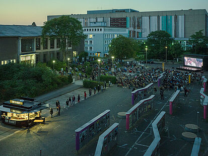 Aus der Vogelperspektive ist der Innenhof der Stasi-Zentrale zu sehen. Zahlreiche Menschen sitzen vor der Kinoleinwand. Im Vordergrund sind die Schautafeln der Open-Air-Ausstellung "Revolution und Mauerfall" zu erkennen.