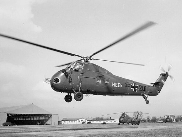 Ein Hubschrauber vom Typ Sikorsky H34G der Heeresflieger schwebt über einem Flugfeld. Im Hintergrund sind weiterere Hubschrauber und ein Hangar zu sehen.