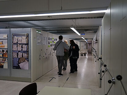 Zwei Besucherinnen und ein Besucher laufen durch das Magazin des Stasi-Unterlagen-Archivs Halle. Links und rechts sind Aktenschränke zu sehen.