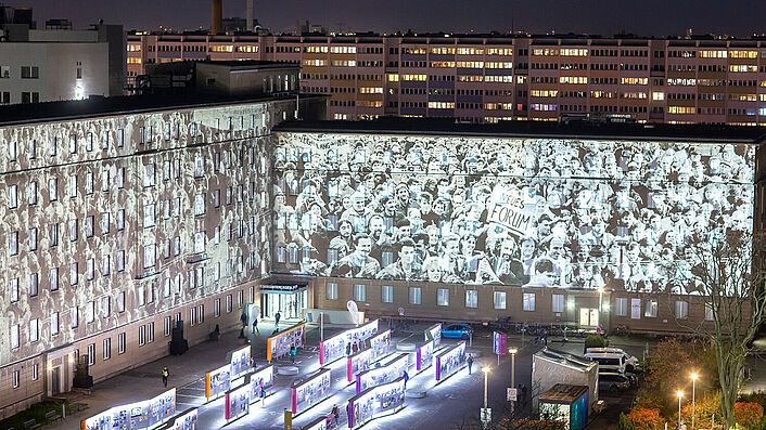 Die Außenfassade von 'Haus 7' auf dem Gelände der ehemaligen Stasi-Zentrale in Berlin-Lichtenberg wird von einem Projektor beleuchtet. Zu sehen sind historische Szenen aus der Zeit des Mauerfalls.