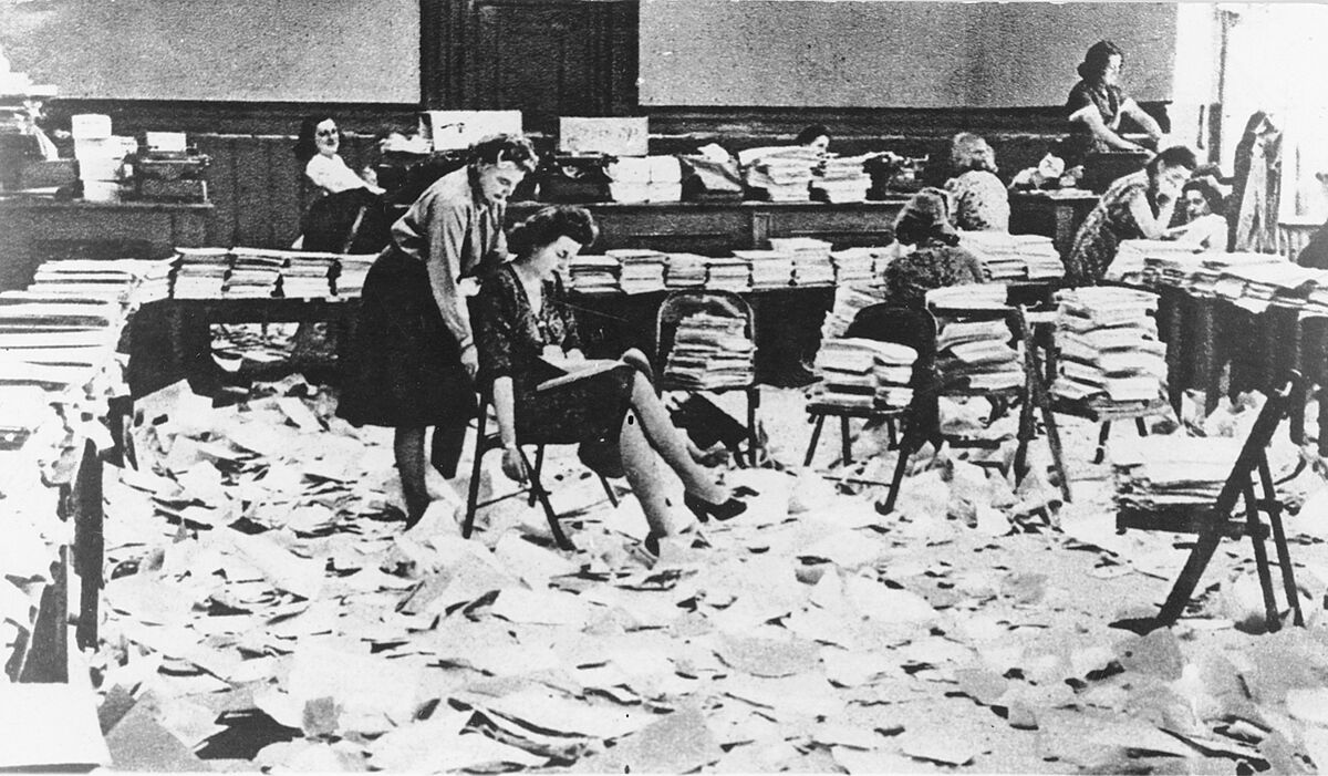 Mehrere Frauen sitzen in einem mit Papieren übersäten Büro und stellen Druckschriften her.