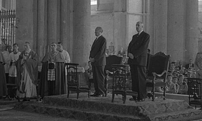 Konrad Adenauer und Charles de Gaulle stehen nebeneinander in einer Kathedrale. Neben ihnen sind mehrere Geistliche, im Hintergrund große Kirchenpfeiler und -fenster zu sehen.