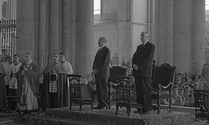 Konrad Adenauer und Charles de Gaulle stehen nebeneinander in einer Kathedrale. Neben ihnen sind mehrere Geistliche, im Hintergrund große Kirchenpfeiler und -fenster zu sehen.