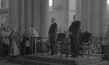 Konrad Adenauer und Charles de Gaulle stehen nebeneinander in einer Kathedrale. Neben ihnen sind mehrere Geistliche, im Hintergrund große Kirchenpfeiler und -fenster zu sehen.