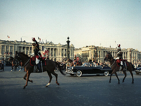 Adenauer und de Gaulle fahren in einer schwarzen Limousine ohne Verdeck vorbei an prachtvollen Gebäuden. Begleitet wird der Wagen von mehreren Reitern in historischen Uniformen. Am Straßenrand stehen Schaulustige.