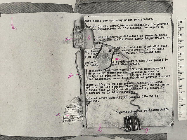 Schwarz-Weiß-Fotografie vom Inneren einer Paketbombe mit Draht und Kabel, mit per Kleinbuchstaben gekennzeichneten Bestandteilen sowie einem eingeklebten Bekennerschreiben in Maschinentext, daneben eine Maßstabsanzeige